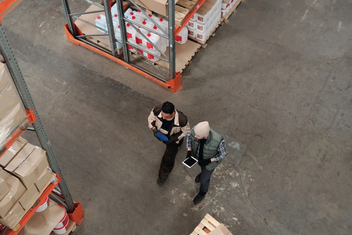 Blick von oben auf zwei Lagerarbeiter mit Tablet zwischen hohen Palettenregalen in einer Halle