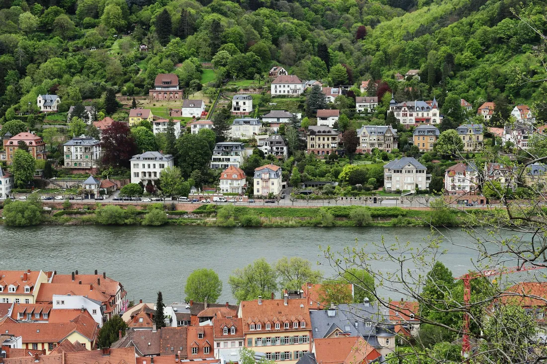 Villen am bewaldeten Hang über einem Fluss, darunter Altstadtdächer mit roten Ziegeln